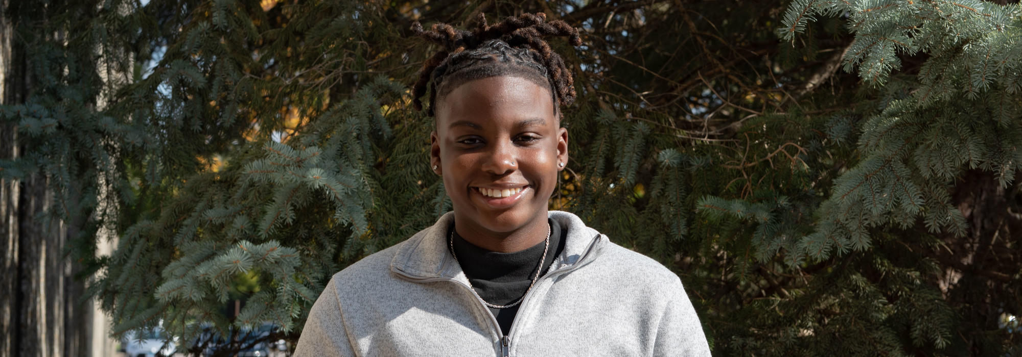 Portrait of Shardae Spruille smiling outdoors in front of evergreen trees.