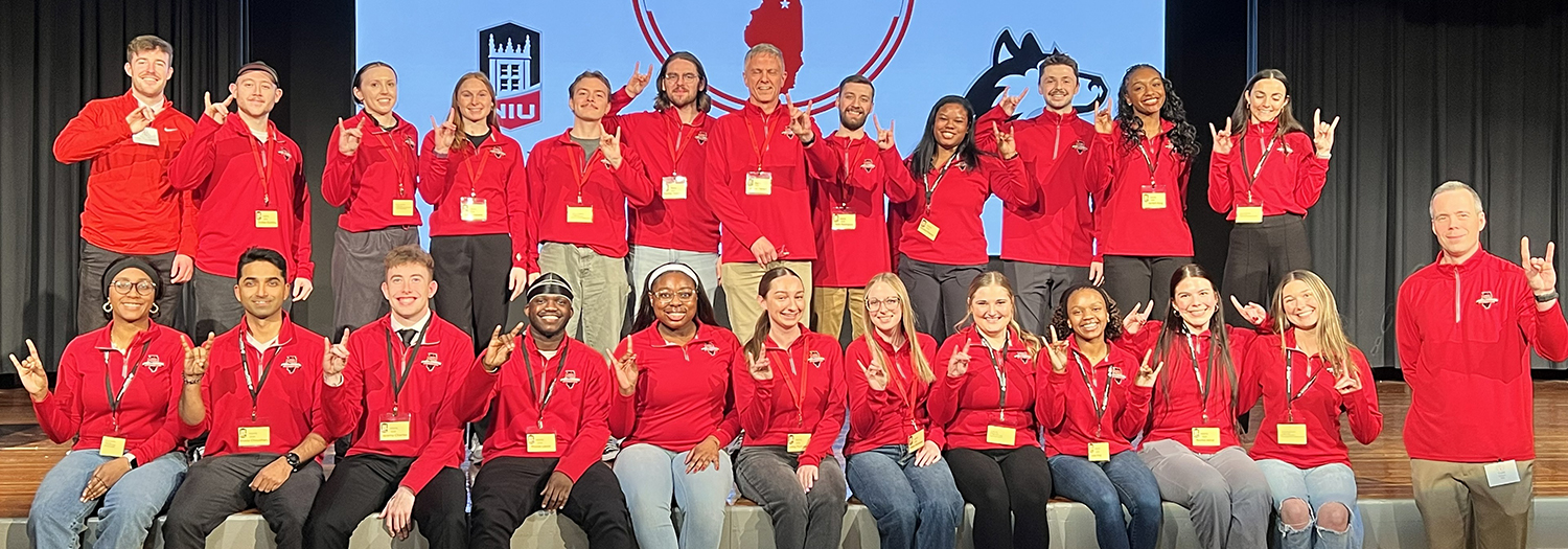 MSEPS conference attendees in matching red NIU shirts pose on a stage and make the NIU hand sign.